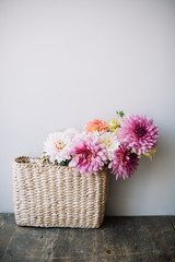Beautiful wicker hand bag with blossoming pink Dahlia flower bouquet in it, bag standing on the wooden table on the grey wall background