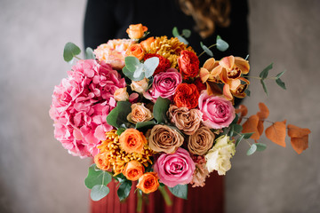 Very nice young woman holding big beautiful blossoming bouquet of fresh hydrangea, roses, carnations, chrysanthemums, cymbidium orchids, eustoma, eucalyptus flowers in pink, red and orange colors