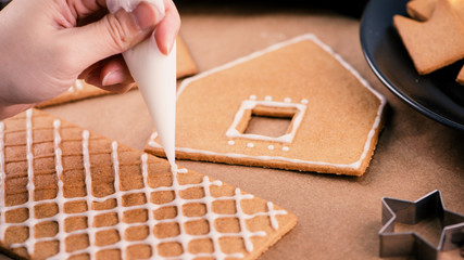Woman is decorating gingerbread cookies house with white frosting icing cream topping on wooden table background, baking paper in kitchen, close up, macro.