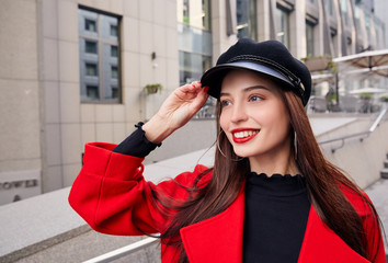 Ravishing luscious delightsome girl in red overcoat holds her kepi by hand, looks into the distance and smiles