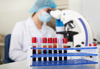 Blurred woman in mask looking in microscope for analyzing blood samples