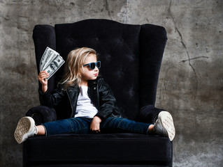 Rich kid girl millionaire sitting in luxury armchair with a bundle of money dollars cash bills looking aside