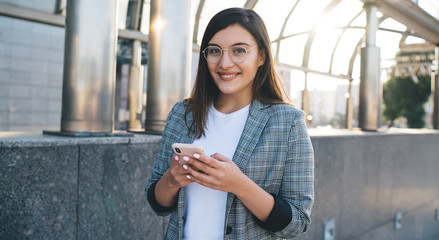 Half length portrait of cheerful caucasian woman in spectacles sending text messages  while strolling on street, positive female blogger looking at camera holding smartphone for chatting online