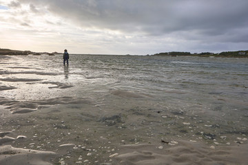 Lonely boy in rainwear standing in the shallow water of the sea at low tide, view to the coast,...