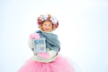 Very sweet beautiful little smiling girl child in pink skirt and mittens, white pullover, gray scarf and floral wreath with old fashioned lantern in hands. Snow is around. Space for text