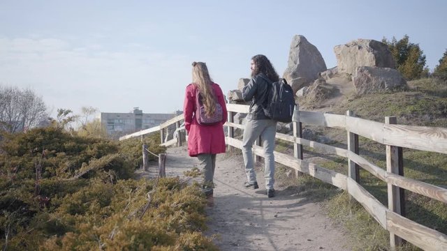 Camera following Middle Eastern man and Caucasian woman walking along wooden fence outdoors. Positive hippie couple dating in autumn park. Togetherness, leisure, resting.