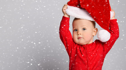 Baby boy toddler in red sweater bodysuit costume playing putting on red christmas cap under the snow on white