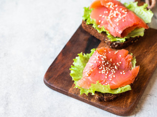 Two toasts with salmon slices, sesame and lettuce on wood board on grey background.