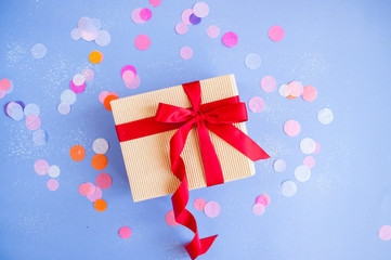 Flat lay of giftbox tied with bright red color ribbon on purple background with sparklets and confetti.