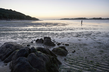Low tide at a beach in Brittany in the evening, lonely boy walking in the shallow water, rocks in the foreground, silent dusk mood