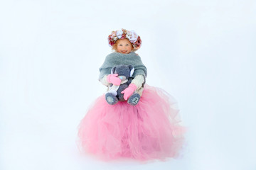 Fototapeta premium Very sweet beautiful little smiling girl child in pink skirt and mittens, white pullover, gray scarf and floral wreath with old taddy bear in hands. Snow around. Space for text