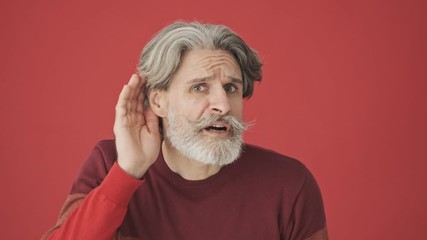 An elderly gray-haired bearded man in a red sweater trying to listen the voice while putting his hand to his ear isolated over red wall