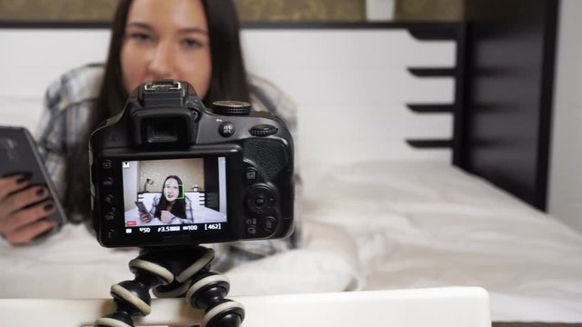 A young girl blogger sits by the window with a camera and records video for the audience. She manages makeup and talks about the process. She actively tells her thoughts to the camera.