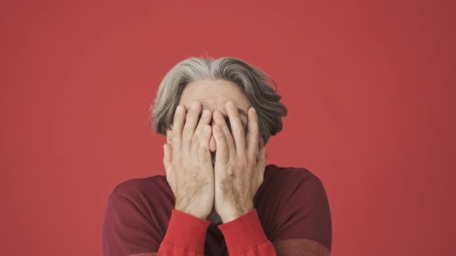
Shocked gray-haired bearded man in the red sweater clutching his head while looking to the camera isolated over red wall