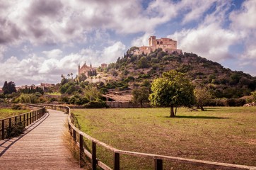 Fototapeta na wymiar Parish church and Pilgrimage church of Sant Salvador Arta, located on a hill with fields and a wooden walkway in the foreground, daytime with blue sky and puffy white clouds, Mallorca, Spain.