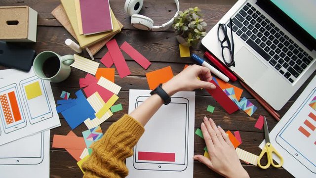 Flat lay of unrecognizable app developer sitting at wooden desk and designing telephone app using layouts and set of colorful pieces of paper
