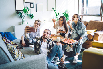 Naklejka premium Friends sitting comfortably on floor eating pizza at table and taking selfie on smartphone