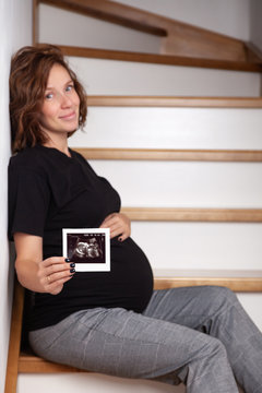 Image beautiful pregnant smiling caucasian curly woman sitting on wooden stairs and posing while showing ultrasound scans on her tummy on white background. Looking at the camera