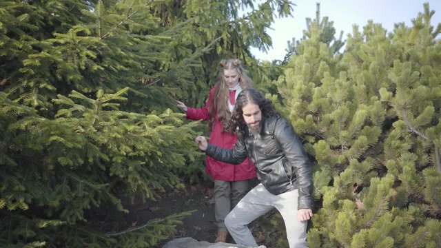 Positive hippie man showing up from trees and leading young beautiful blond woman. Happy young multiracial couple dating in autumn forest. Cheerful people resting outdoors.