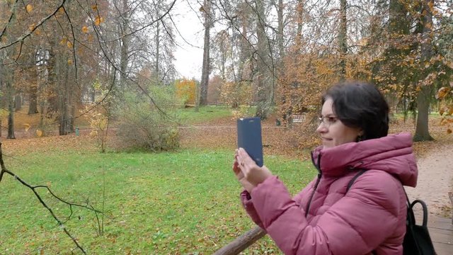 Cloudy autumn day Park Caucasian a middle aged woman in a Burgundy down jacket glasses a black backpack behind her back takes pictures on a smartphone nature and what he says Medium shot