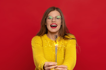 excited girl in eyeglasses holding sparklers, wears yellow sweater, looks with happiness, expresses good emotions, isolated on red background. Christmas concept
