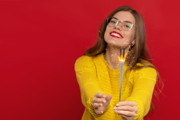 Obraz na płótnie Canvas selective focus of happy girl in eyeglasses holding sparklers, wears yellow sweater, isolated on red background, expresses good emotions. Christmas concept