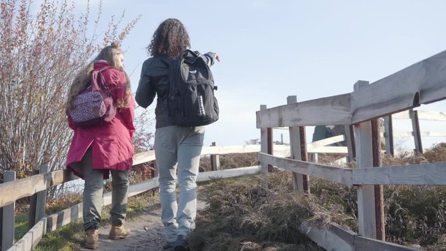 Back view of young multiracial couple with backpacks going up along the fence in autumn park. Adult boyfriend and girlfriend strolling outdoors. Carefree people dating.