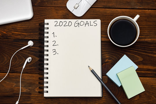 2020 goals text on notepad, chek list. Top view of headphones, mouse, coffee and notebook on wooden background. Flat lay.