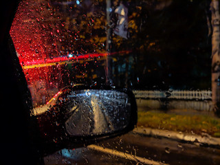 Amazing atmosphere during night while driving on a rainy street with closeup of red car street lights and drops of rain in focus on a car window glass with side rear view mirror