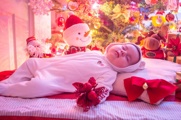 Fototapeta na wymiar A sleeping baby on a cushion dressed in white, surrounded by dolls, bow and red flower, lights and Christmas decorations