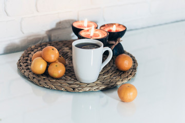 Burning spa aroma candles in coconut shell, cup of tea, tangerines on wicker stand on a glass white table, cozy home interior background.