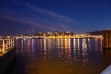 Fototapeta premium Romantyczna jesienna wieczorna panorama nad wodami kanału holenderskiego miasta metropolitalnego Amsterdam, wśród świateł i iluminacji budynków i drapaczy chmur
