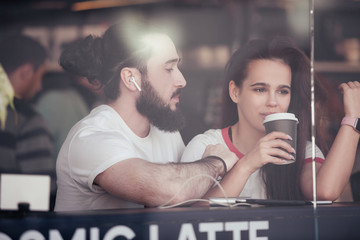View through the glass nice young couple in love chatting and looking at each other sit at a table in a cafe with a cup of coffee and a smartphone. Concept free high-speed wi-fi, live communication