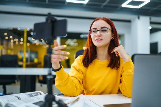 Female blogger with laptop recording video while sitting at office and talking  about startup small business. Teenager student in a yellow sweater having fun vlogging live feeds on social media.
