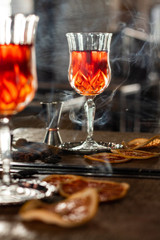 cocktail in a flute, reflected in the mirror on a wooden table, with cinnamon, dried orange slices and cardamom seeds. vertical visual with incense smoke background