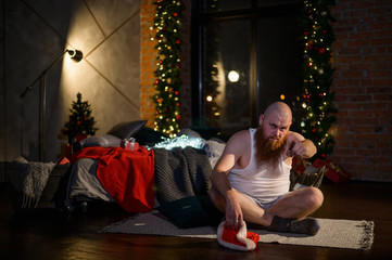 Obraz na płótnie Canvas Humorous image of a man without pants in front of Christmas. Bald Santa Claus with a red beard unpacks a gift. A parody of a glamorous women's New Year photo shoot. Joke.