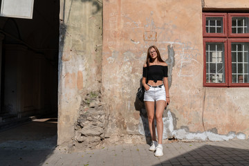 Young attractive brunette girl in glasses, dressed in white shorts and black top, standing near the wall of the old house and looking at the camera