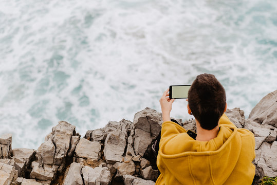 Back view of short haired unrecognizable woman in yellow sweatshirt sitting on rocky seashore above foamy wavy ocean and taking selfie on mobile phone on gray cloudy day