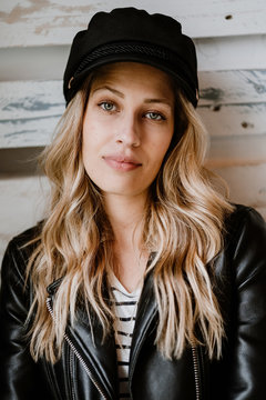 Stylish beautiful confident woman in leather jacket and trendy black cap looking at camera on wooden wall