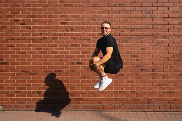 Young man in sunglasses, dressed in black casual clothes jump and hugged his knees with his hands, red brick wall on the background