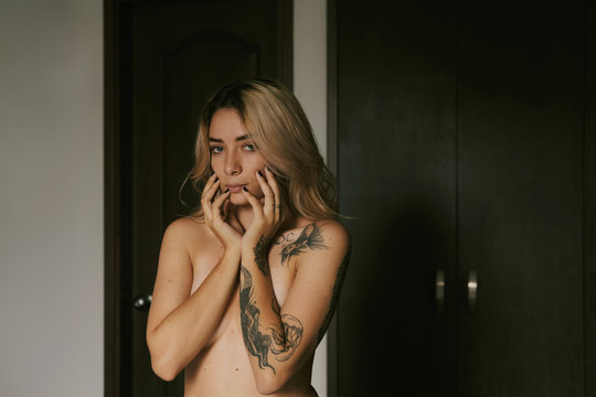 Sexy seductive topless fair hared female with tattoos on shoulder and arm covering breast and touching face 