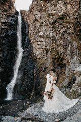 Fototapeta na wymiar noise effect, selective focus: incredibly enamored brides hugging, kissing and posing for a photo on the incredible rocky mountains background with a big waterfall