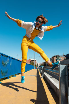 From below cheerful courageous teenager in sunglasses and trendy yellow clothes jumping high with mouth open and arms raised on street bridge against blurred urban view in sunny day