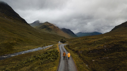 From above calm couple looking away and enjoying view while standing on empty road and holding hands near mountain river in green lush grassland against blurred misty valley in overcast weather in Scotland