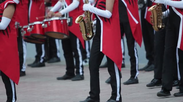 several saxophone hispanic players in a marching band moving their feet from left to right wearing black pants with white stripes, red jackets and white gloves