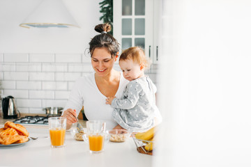Obraz na płótnie Canvas Young woman mom with baby girl on hands cooking breakfast on bright kitchen at home