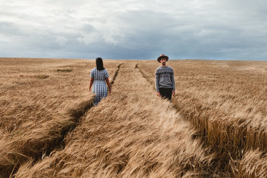 Woman in checked dress and man in sweater and hat walking in opposite directions on parallel footpaths among boundless brown Scottish field with cloudy sky on background 