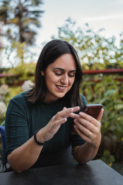 Young brunette in casual clothes browsing internet on smartphone and laughing at funny photo while resting on terrace with green plants on background