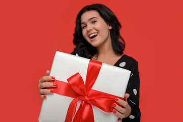 Brunette woman in a black dress smiling holding Christmas giftbox of white color with red ribbon.