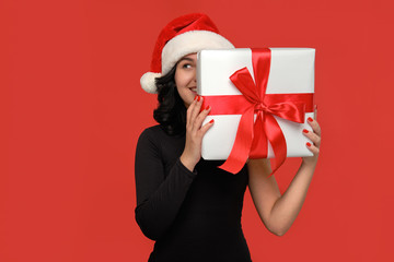 Woman in a black dress and Santa hat hiding behind Christmas giftbox of white color with red ribbon.